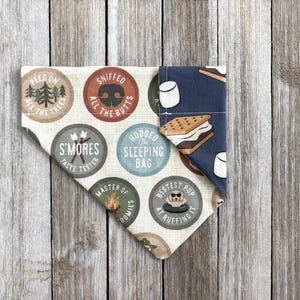 May include: A dog bandana with a camping theme. The bandana features various circular designs with text like "Peed on all the trees" and "S'mores taste tester." The other side has a blue background with s'mores and marshmallow graphics.