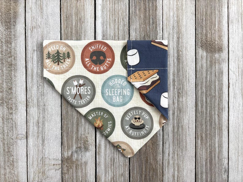 May include: A dog bandana with a camping theme. The bandana features various circular designs with text like "Peed on all the trees" and "S'mores taste tester." The other side has a blue background with s'mores and marshmallow graphics.