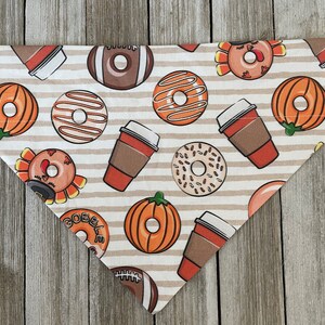 May include: A beige and brown dog bandana with a Thanksgiving theme. The bandana features images of pumpkins, donuts, coffee cups, and turkeys. The text "Gobble" is printed on the bandana.