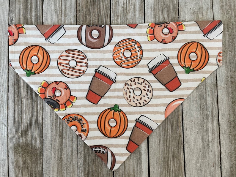 May include: A beige and brown dog bandana with a Thanksgiving theme. The bandana features images of pumpkins, donuts, coffee cups, and turkeys. The text "Gobble" is printed on the bandana.