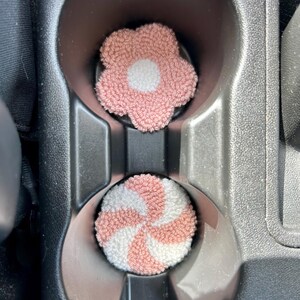 May include: Two car coasters. One is a pink flower shape with a white center. The other is a round coaster with a pink and white spiral design. Both are inside a car's cup holders.