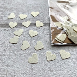 Pode incluir: Um saco de confete de papel branco em forma de cora&ccedil;&atilde;o. Os cora&ccedil;&otilde;es est&atilde;o espalhados sobre uma superf&iacute;cie branca.