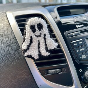 May include: A white and black ghost-shaped car air freshener. The ghost has a smiling face with black eyes and mouth. The air freshener is attached to a car vent, adding a touch of spooky decor.