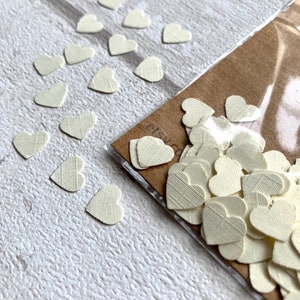 Pode incluir: Um saco de confete de papel branco em forma de cora&ccedil;&atilde;o. Os cora&ccedil;&otilde;es s&atilde;o pequenos e t&ecirc;m uma superf&iacute;cie texturizada. Tamb&eacute;m h&aacute; alguns cora&ccedil;&otilde;es espalhados sobre uma superf&iacute;cie branca.