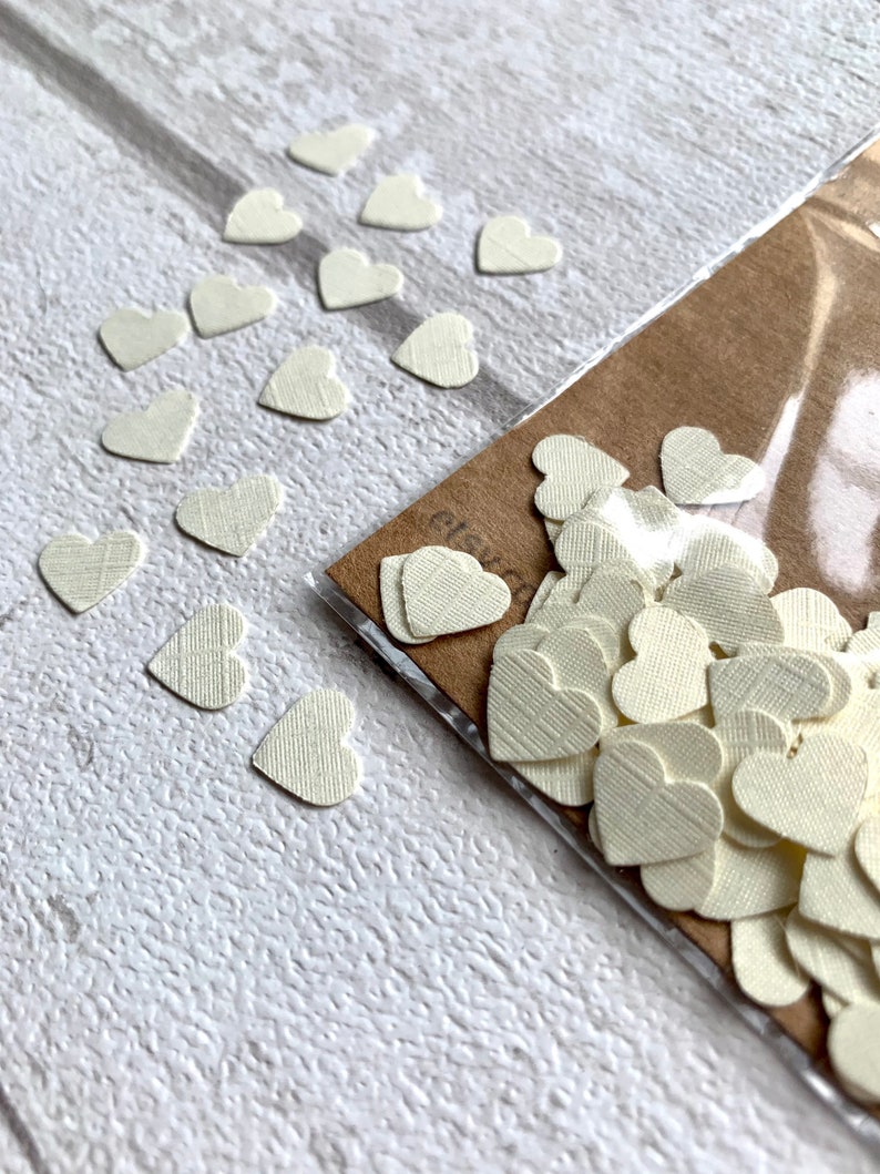 Pode incluir: Um saco de confete de papel branco em forma de cora&ccedil;&atilde;o. Os cora&ccedil;&otilde;es s&atilde;o pequenos e t&ecirc;m uma superf&iacute;cie texturizada. Tamb&eacute;m h&aacute; alguns cora&ccedil;&otilde;es espalhados sobre uma superf&iacute;cie branca.