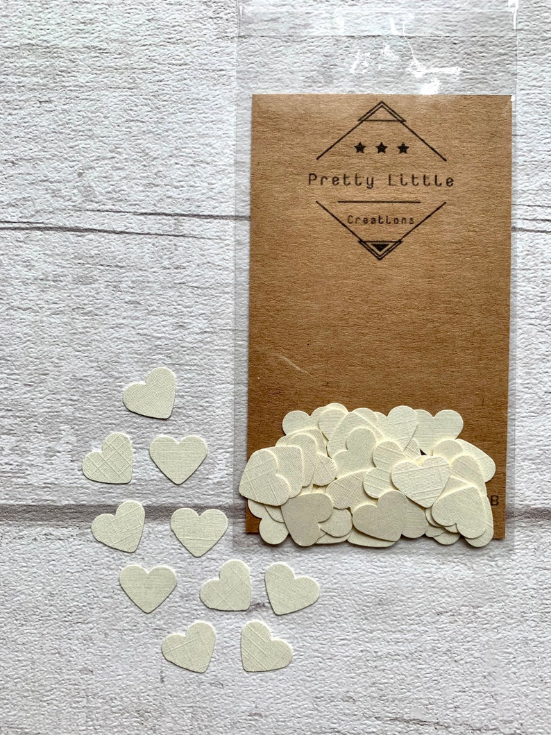Pode incluir: Um saco de confete de papel branco em forma de cora&ccedil;&atilde;o com um cart&atilde;o marrom que diz "Pretty Little Creations" em texto preto.