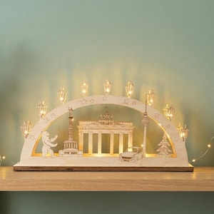 May include: A wooden Christmas decoration with illuminated Berlin landmarks, including the Brandenburg Gate and the TV Tower. The arch-shaped piece is set on a wooden surface with a vase of greenery and folded blankets.