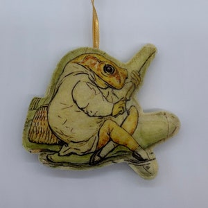 May include: A plush ornament featuring a character from the children's book 'The Tale of Mr. Tod' by Beatrix Potter. The ornament depicts Mr. Tod, a fox, sitting on a log with a fishing rod in his hand. The ornament is made of soft, plush fabric and has a gold ribbon for hanging.