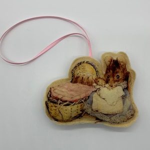 May include: A felt ornament featuring a mouse holding a basket with a baby mouse inside. The ornament is beige with a pink ribbon attached.