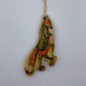 May include: A felt ornament featuring a fox wearing a green jacket and holding a walking stick. The ornament is attached to a gold ribbon.