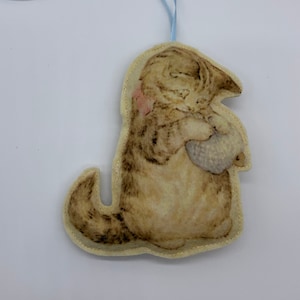 May include: A beige felt ornament of a brown and white kitten holding a small white mouse. The ornament has a light blue ribbon attached for hanging.