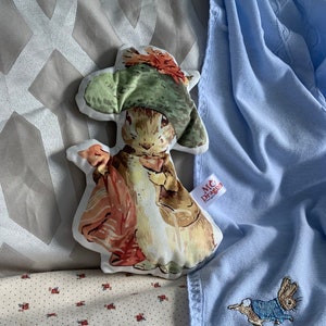 Könnte beinhalten: Eine blaue Babydecke mit einem weißen gestickten Peter-Rabbit-Motiv. Die Decke liegt auf einem cremefarbenen und roten Stoff mit Blumenmuster. Links im Bild befindet sich ein Peter-Rabbit-Plüschtier.