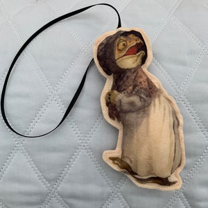 May include: A felt ornament of a frog wearing a bonnet and dress. The frog is brown and green with a white dress and a black bonnet.