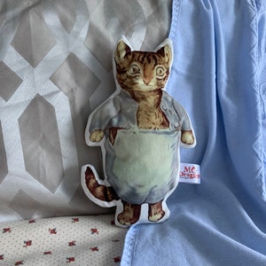 May include: A plush toy of a cat wearing a blue and white outfit. The cat has brown fur and green eyes. The toy is sitting on a blue blanket with a white pattern.