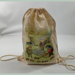 May include: A beige canvas drawstring bag with a printed image of characters from the book "The Wind in the Willows". The characters are Mr. Toad, Ratty, Old Badger, and Mole. The bag has a white drawstring.