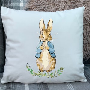 May include: White cushion with a blue background featuring a cartoon illustration of Peter Rabbit wearing a blue jacket and standing on a bed of green leaves.