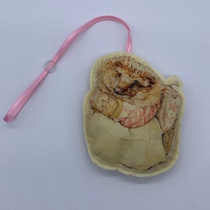 May include: A beige fabric ornament with a pink ribbon loop. The ornament features a printed image of a hedgehog holding a mushroom.