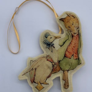 May include: A felt ornament featuring a fox and a duck. The fox is wearing a red vest and green pants. The duck is wearing a blue hat and a yellow ribbon. The ornament is attached to a gold ribbon.
