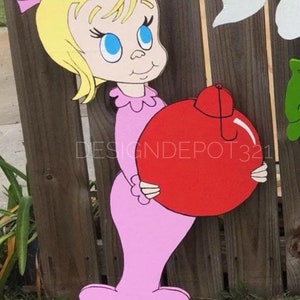 May include: A pink and white cutout of a girl character holding a red ornament with a white top. The character has blonde hair and blue eyes. The cutout is against a wooden fence background.