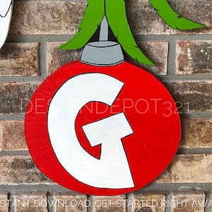Peut inclure: Boule de Noël rouge avec une lettre G blanche dessus. La boule est suspendue à une tige verte et blanche.