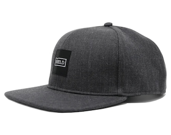 May include: A dark grey, flat-brimmed baseball cap with a black square patch on the front that says "SHIELD" in white letters.