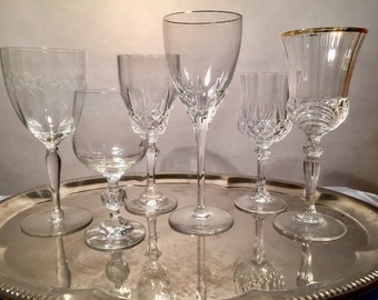 May include: A set of six clear glass drinking glasses of various shapes and sizes, including a wine glass, a champagne flute, a brandy snifter, and a shot glass. The glasses are arranged on a silver tray.