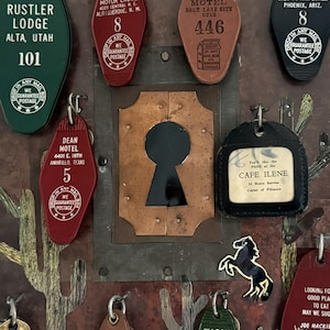 May include: A collection of vintage motel keychains and a keyhole-shaped copper accent. Keychains are various colors, including green, red, and black, with motel names and numbers. The keyhole is surrounded by a metal frame with a desert landscape design.