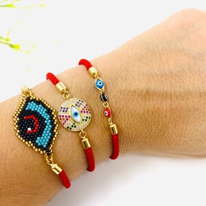 Puede incluir: Tres pulseras de hilo rojo con dijes de ojo turco. Una pulsera presenta un dije de ojo turco grande y redondo con pedrería de colores. Otra pulsera tiene un dije de ojo turco más pequeño, azul y negro, con detalles de cuentas. La tercera pulsera tiene una serie de dijes de ojo turco más pequeños en rojo, negro y azul.