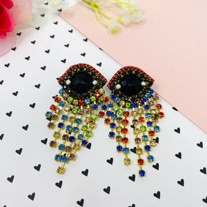 Puede incluir: Un par de pendientes colgantes con un diseño de ojo negro rodeado de piedras de cristal de colores. Los pendientes tienen una larga cadena de piedras de cristal de colores en cascada.