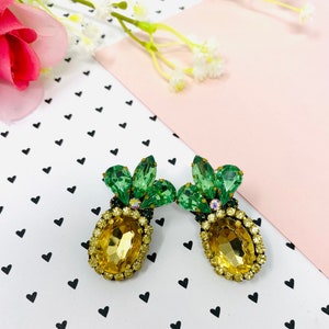 Puede incluir: Un par de pendientes de piña dorados con hojas de cristal verde. Los pendientes están sobre un fondo blanco con corazones negros.