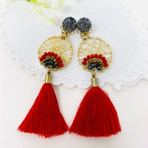 Puede incluir: Pendientes de alambre dorado con borlas rojas. Los pendientes presentan un diseño circular con cuentas rojas y negras, y un perno gris brillante. Las borlas son de un rojo vibrante.