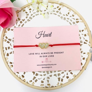 Puede incluir: Pulsera de cuerda roja con un dije de doble corazón dorado con brillantes transparentes. La pulsera está sobre una tarjeta rosa con el texto "Heart" y "Love will always be present in our lives".