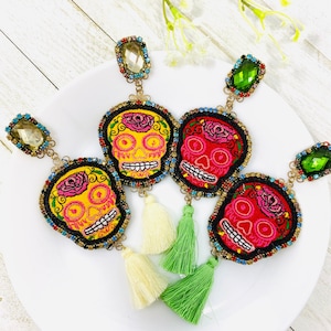 Puede incluir: Cuatro pares de pendientes de calavera coloridos con borlas. Cada pendiente presenta una calavera con detalles bordados y un adorno de cristal. Las calaveras son en tonos de rojo, rosa, amarillo y azul.
