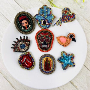 Puede incluir: Una colección de nueve broches de cuentas coloridas con varios diseños, incluyendo un corazón, una calavera, una estrella, un ojo, una mano, un pájaro y un retrato de Frida Kahlo.