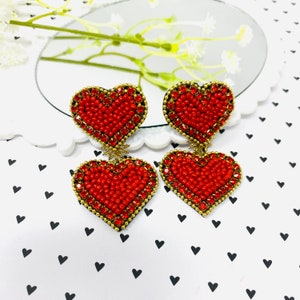 Puede incluir: Un par de pendientes colgantes de corazón rojo con detalles dorados. Los pendientes están hechos de pequeñas cuentas rojas y tienen un acabado brillante.