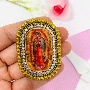 Puede incluir: Broche de forma ovalada con un marco de metal dorado y una imagen colorida de la Virgen María. El marco está adornado con diamantes de imitación transparentes.