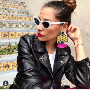 May include: A woman wearing a black leather jacket with a white t-shirt underneath. She is wearing white cat-eye sunglasses and large pink and yellow tassel earrings with blue accents.