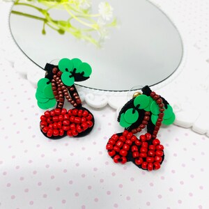 Puede incluir: Un par de pendientes de cereza rojos y verdes. Las cerezas están hechas de cuentas y las hojas están hechas de lentejuelas verdes.