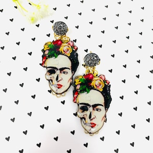 Puede incluir: Un par de pendientes colgantes con un retrato colorido de Frida Kahlo con un diseño de calavera. Los pendientes tienen un acabado dorado y están adornados con diamantes de imitación.