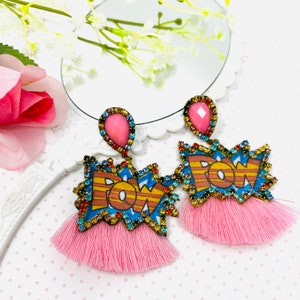 Puede incluir: Un par de pendientes de borla rosa con un diseño "POW" colorido y detalles de cristal rosa.