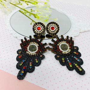 Puede incluir: Un par de pendientes negros en forma de ojo con cuentas de colores y un centro redondo como un espejo. Los pendientes tienen un diseño colgante con un borde festoneado.