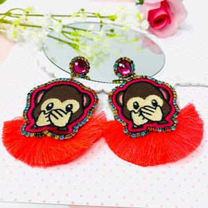Puede incluir: Un par de pendientes de borla naranja brillante con un diseño de cara de mono. El mono se está tapando la boca con la mano, y los pendientes están decorados con brillantes de colores.