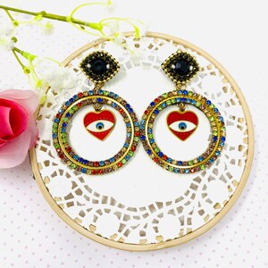 Puede incluir: Pendientes de aro dorados con un dije de corazón rojo con un ojo azul mal de ojo. Los pendientes están decorados con piedras de cristal de colores.