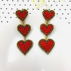 Puede incluir: Un par de pendientes colgantes con tres colgantes en forma de corazón rojo. Cada corazón está hecho de pequeñas cuentas rojas y tiene un contorno dorado. Los corazones están conectados por una cadena dorada.