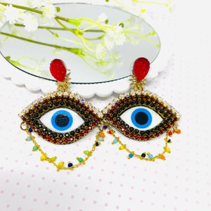 Puede incluir: Un par de pendientes con forma de ojo. Los pendientes presentan un iris blanco con una pupila azul, rodeado por un borde negro y rojo enjoyado. Una gema roja texturizada remata cada pendiente, con detalles dorados y cadenas colgantes con cuentas de colores.