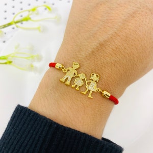 Puede incluir: Pulsera de cuerda roja con un dije de oro que representa a una familia de tres figuras de palo. Las figuras se toman de la mano y tienen pequeñas piedras brillantes para los detalles.