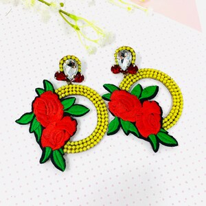 Op de afbeelding: Een paar grote statement-oorbellen met een gele kralencirkel met een rood roos- en groen bladontwerp. Elke oorbel heeft een heldere kristallen strass in het midden.