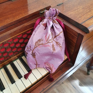 May include: A purple satin drawstring bag with a floral pattern and burgundy tassels. The bag is sitting on top of a wooden piano.