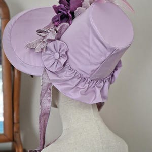 May include: A lavender bonnet-style hat with a cylindrical crown and a wide brim. The hat is adorned with purple fabric flowers, ribbons, and a ruffled edge. A long, dotted ribbon hangs from the side.