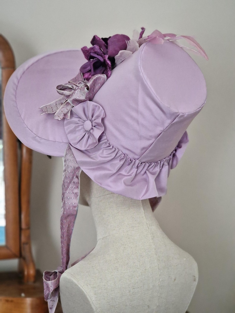 May include: A lavender bonnet-style hat with a cylindrical crown and a wide brim. The hat is adorned with purple fabric flowers, ribbons, and a ruffled edge. A long, dotted ribbon hangs from the side.